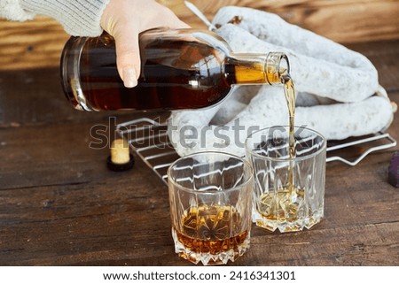 Women's hands pour cognac into two glasses. In the background there is a sausage with white mold, elite varieties.