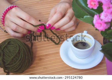 Women's hands with purple manicure are knitted metal spokes of a wooden table. On the table is a cup of coffee, flower pot and a green ball of wool yarn.