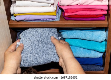 Women's Hands Put Clean Clothes On A Shelf In A Closet At Home. A Woman Puts Baby Clothes On A Bookcase. House Cleaning, Order, Accuracy In The Wardrobe