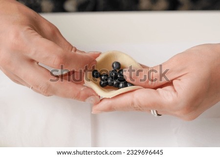 Women's hands sculpt Ukrainian dumplings with blueberries on a light background. A close-up of the preparation of a traditional dessert made of dough and berries