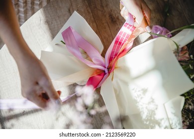 Women's Hands Tie With A Pink Ribbon A Bouquet Of Flowers, Wrapped In Paper, Lying On A Wooden Table