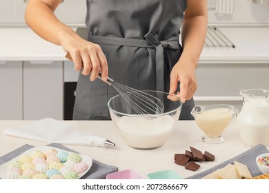 Women's hands whisk cream, cream, mousse, dessert in a transparent bowl against the background of a white kitchen. Grey apron. Marshmallows, cream, milk, chocolate. Side view. Confectionery.