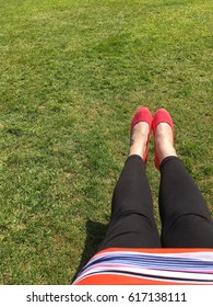 Women's Legs In Black Leggings And Colourful Striped Dress Wearing Pink Shoes Stretched Out Over Grass On Sunny Day. Female Body POV. Chilling Out Concept.