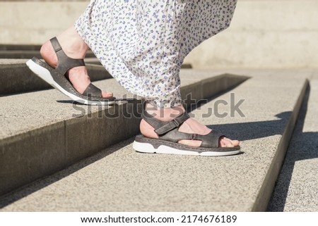 Womens legs in open leather sandals with flat soles go down the stairs. A woman in a light summer dress walks down the stairs.