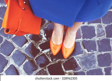 Women's legs in orange shoes. Bright orange shoes and bag. Blue coat, orange classic ladies shoes and tote bag. Rainy day. Street fashion. Street style. Business casual look. Autumn outfit.