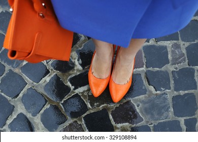 Women's legs in orange shoes. Bright orange shoes and bag. Blue coat, orange classic ladies shoes and tote bag. Rainy day. Street fashion. Street style. Business casual look. Autumn outfit.
