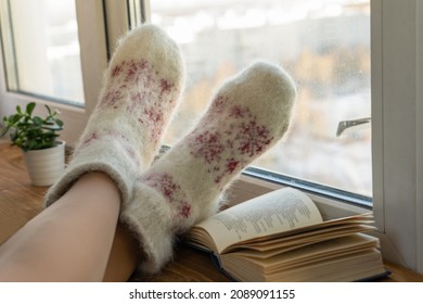 Women's Legs In Warm Woolen White Socks Snuggle Up With A Book On The Windowsill Looking Out The Window In The Cold Winter