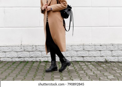 Women's legs. Woman wearing long beige coat, black boots and leather backpack walking through the city streets. Trendy casual outfit. Details of everyday look. Street fashion.