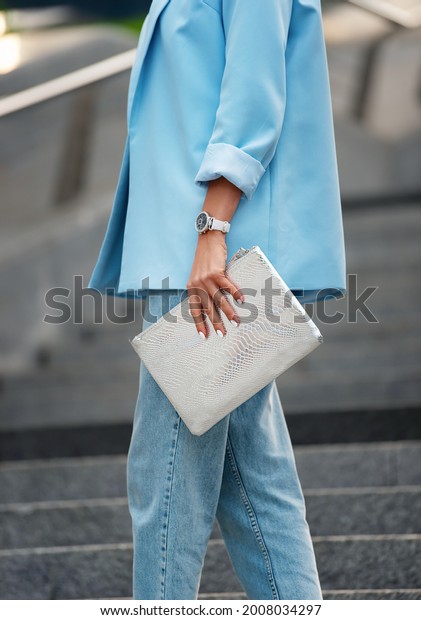 Women\'s silver clutch bag\
with snakeskin texture. A fragment of the body of a model in a\
summer blue blazer with rolled-up sleeves and blue jeans. Business\
style clothing.