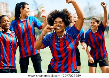 Women's soccer team celebrating victory on a playing field. Focus is on African American player biting her gold medal. 