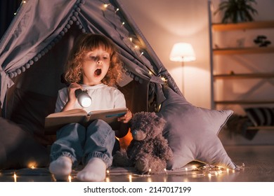 Wondered by book. Using flashlight. Cute little girl is in the tent in domestic room.