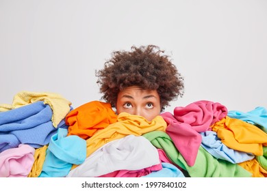 Wondered curly haired ethnic woman focused above surrounded by multicolored laundry cluttered with clothes collects clothing for recycling isolated over white background. Organize your closet - Powered by Shutterstock