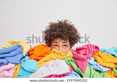 Wondered curly haired ethnic woman focused above surrounded by multicolored laundry cluttered with clothes collects clothing for recycling isolated over white background. Organize your closet