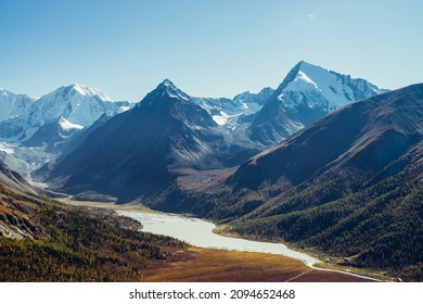 Wonderful alpine landscape with mountain lake and mountain river in valley with forest in autumn colors on background of snowy mountains silhouettes under blue sky. Beautiful mountain valley in autumn