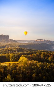 Wonderful Atmosphere At The Morning In The Elbe Sandstone Mountains. The Sun Is Rising And The Light Is Coming. Great Landscape With Forest An Mointain. A Hot Air Balon Is Flying.