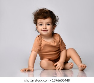 Wonderful curly kid tries to crawl, sat down to rest. he is dressed in a beige bodysuit on a gray background - Shutterstock ID 1896603448