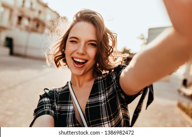 Wonderful Curly Woman With Dark Eyes Playfully Posing On The Street. Outdoor Photo Of Inspired Young Lady Making Selfie.