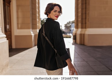 Wonderful girl in black jacket with dark handbag smiling at street. Short-haired woman in eyeglasses with red lips posing outside..