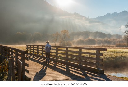 Wonderful Misty Morning At Alpine Mountais. Elderly Woman with Nordic Sticks, Hiking in the Foggy Meadow under Sunlight. Active and Healthy Lifestyle. Nordic Walking Exercise Adventure Hiking Concept.