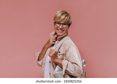 Wonderful Old Woman With Blonde Hair And Stylish Glasses In Beige Jacket And Light T-shirt Smiling And Posing With Bag On Pink Background..