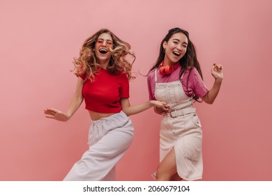 Wonderful Young Racial Girls Are Dancing To Beat Of Music On Pink Background In Studio. Pretty Women In Colorful Casual Clothes Are Smiling Broadly Holding Hands. Merry Holiday Concept