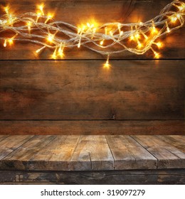 Wood Board Table In Front Of Christmas Warm Gold Garland Lights On Wooden Rustic Background. Filtered Image. Selective Focus
