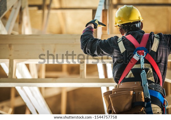 Wood\
Construction Worker. Caucasian Builder Wearing Safety Harness and\
the Wooden House Structure. Industrial\
Theme.