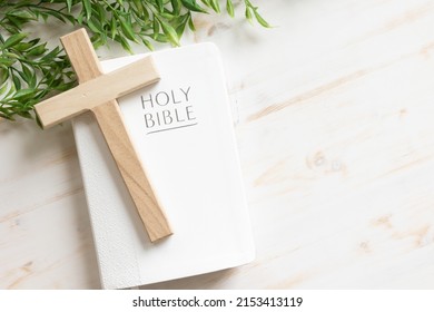 Wood cross laying on a closed white Christian bible with a green vine border all on a white wood background with copy space