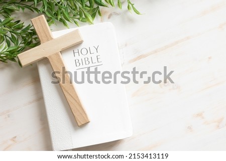 Wood cross laying on a closed white Christian bible with a green vine border all on a white wood background with copy space
