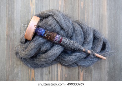 Wood Drop Spindle With Handmade Yarn Made From The Wool Of A Sheep On A Wood Table On Top Of A Pile Of Raw Gray Wool Roving