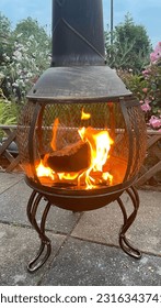 A wood fire burning in a garden metal chimnea on a patio with a fence and garden plants in background