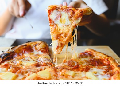 Wood Fire Cheesy Pizza Slice In Hand. Traditional Hawaiian And Salami Pizza Savory Dish Flavour Of Origin Italian Restaurant. Wood-Fired Oven Pizza With People Holding And Cutting Knife Background