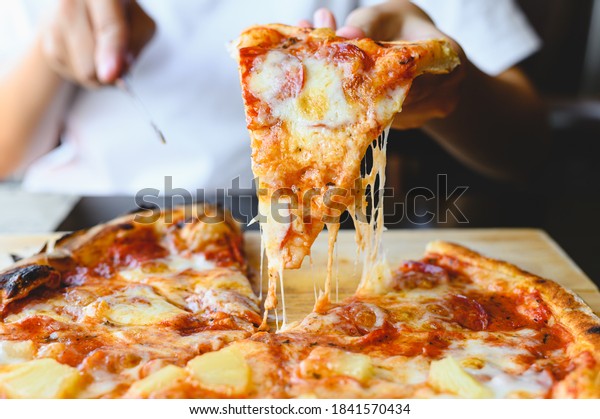 Wood fire cheesy pizza slice in hand.\
Traditional Hawaiian and Salami pizza savory dish flavour of origin\
Italian restaurant. Wood-Fired Oven Pizza with people holding and\
cutting knife background