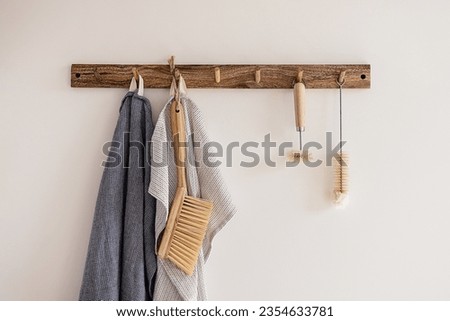 Wood hook rack with tea towels, broom and dish bottle brush hanging on a white wall on the modern style kitchen background. Smart organisation storage ideas. Eco friendly life. Natural materials.