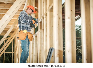 Wood House Frame Construction and the Contractor Worker in His 30s with Drill Driver Attaching Wooden Frame Elements. Industrial Theme.