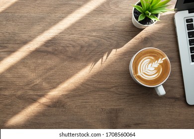 Wood Office Desk Table With Cup Of Latte Coffee And Laptop Computer With Sun Light. Top View With Copy Space, Flat Lay. Working From Home In The Morning Concept.