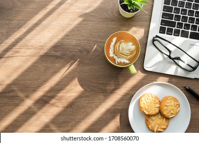 Wood office desk table with dish of cookies, cup of latte coffee and supplies. Top view with copy space, flat lay. Working from home in the morning concept.