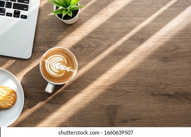 Schreibtisch aus Holz mit Laptop-Computer, Mandelkekse und eine Tasse latten Kaffee mit Morgensonnenlicht. Frühstück Snack Konzept. Draufsicht mit Kopienraum, flach gelegt.