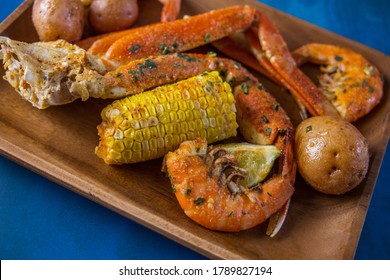 A Wood Platter Of Boiled Southern Garlic Seafood Including Shrimp, Crab Legs, Corn On The Cob, And New Potatoes On A Bright Blue Surface