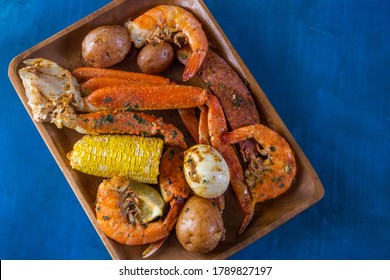 A Wood Platter Of Boiled Southern Garlic Seafood Including Shrimp, Crab Legs, Corn On The Cob, And New Potatoes On A Bright Blue Surface