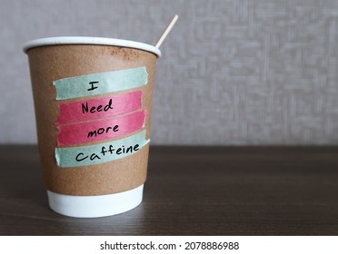 Wood stirrer in paper coffee cup with text written I NEED MORE CAFFEINE , concept of caffeine addiction, daily coffee intake to boost energy and wake up in the morning,
