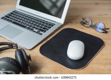 Wood Work Surface With A Smartphone, Headphone, A Pair Of Glasses, A Laptop, A White Wireless Mouse And A Black Leather Mouse Pad.