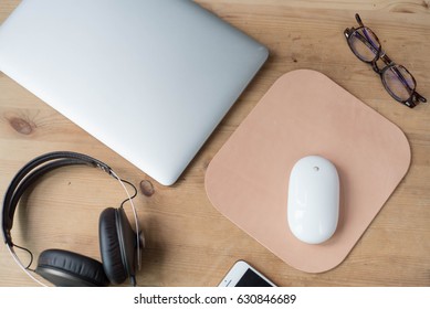 Wood Work Surface With A Smartphone, Headphone, A Pair Of Glasses, A Laptop, A White Wireless Mouse And A Leather Mouse Pad.