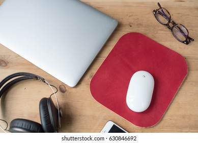 Wood Work Surface With A Smartphone, Headphone, A Pair Of Glasses, A Laptop, A White Wireless Mouse And A Red Leather Mouse Pad.