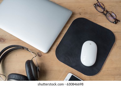 Wood Work Surface With A Smartphone, Headphone, A Pair Of Glasses, A Laptop, A White Wireless Mouse And A Black Leather Mouse Pad.