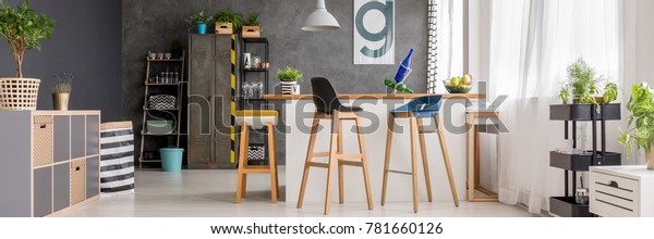 Wooden bar stools\
at dining room interior with bottles in open kitchen with plants\
and poster on concrete\
wall