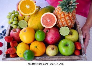 Wooden Basket On White Table Full Of Fresh Colorful Fruits. Mature Woman Holding Half Orange Fruit. Healthy Eating And Lifestyle