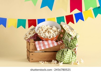 Wooden basket with tasty popcorn, bundle bag and flags for Festa Junina celebration on color background