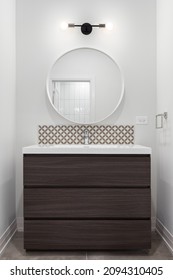 A Wooden Bathroom Vanity Cabinet With Marble Countertop, Tiled Backsplash, And A Light Over A Circular Mirror.