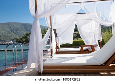 Wooden beach canopies covered by fabric on the beach of the Mediterranean Sea. White curtain being blow by wind. Comfortable chaise lounges on the zone of relaxation next to the pool, romantic place.